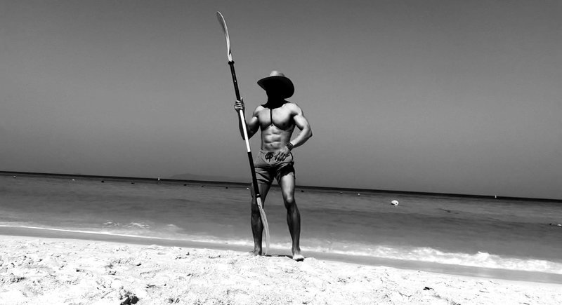 Masked figure standing on a beach holding a paddle vertically.