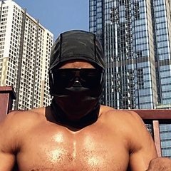 Portrait of OYOTTA wearing a dark head covering and sunglasses, with high-rise buildings behind.