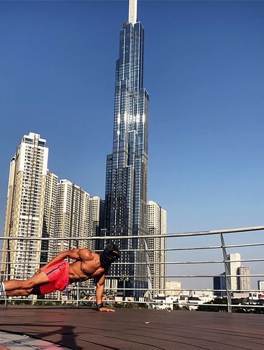 An athlete holding a one-armed side plank on a wooden rooftop deck, with a tall glass tower rising behind.