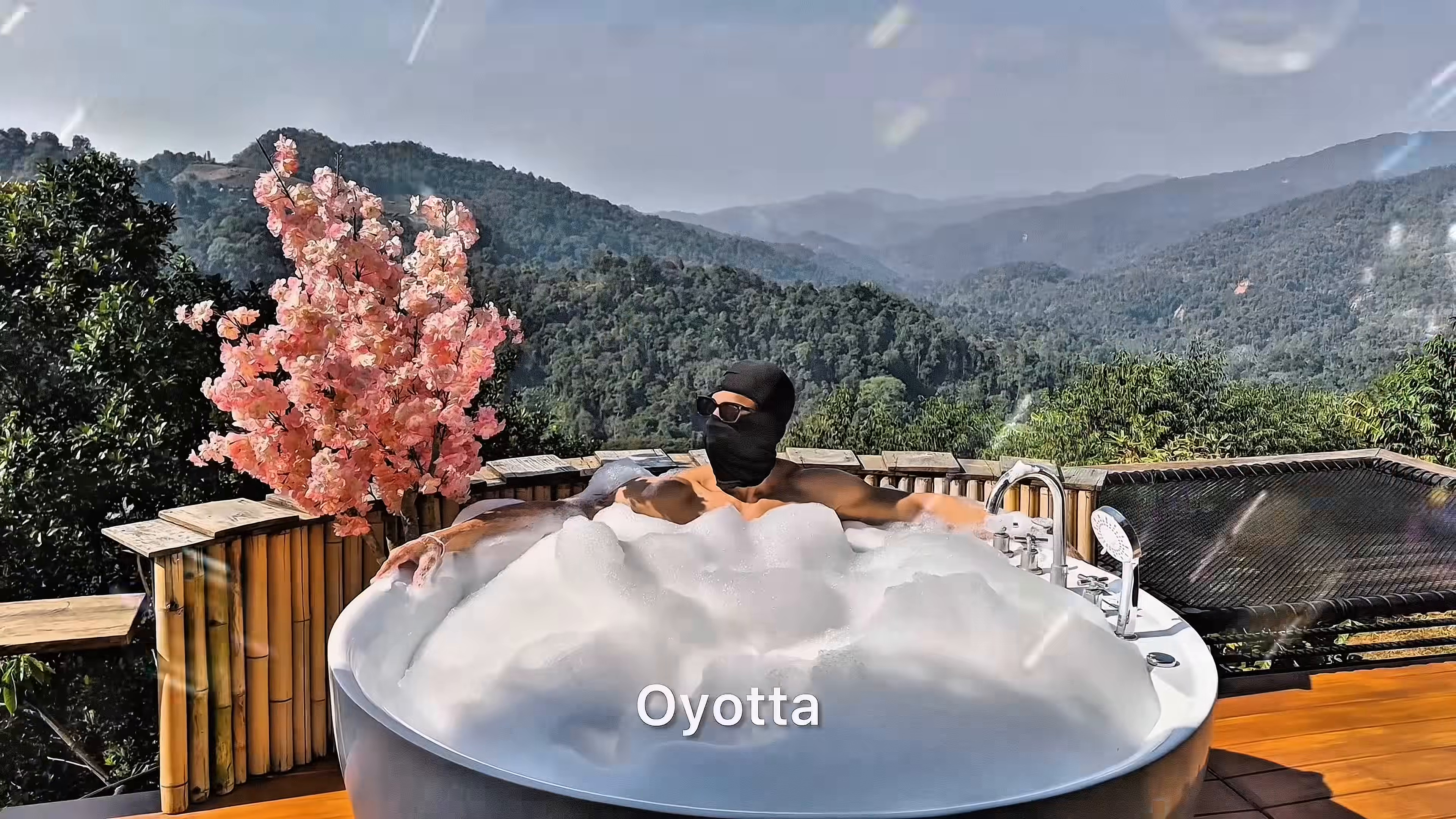 A masked figure sits in a foaming pool on a wooden terrace, with a flowering tree and layered green mountains behind.