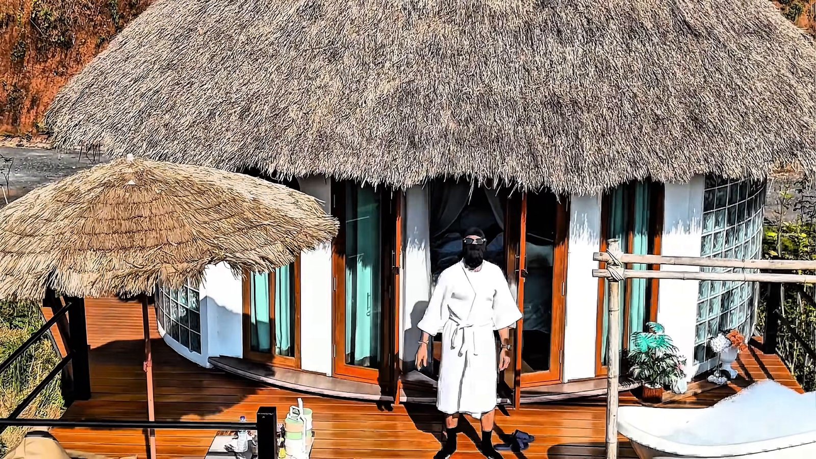 The masked figure faces the camera in a white robe on a wooden deck, framed by an open doorway, a thatched roof and a shaded outdoor bath.